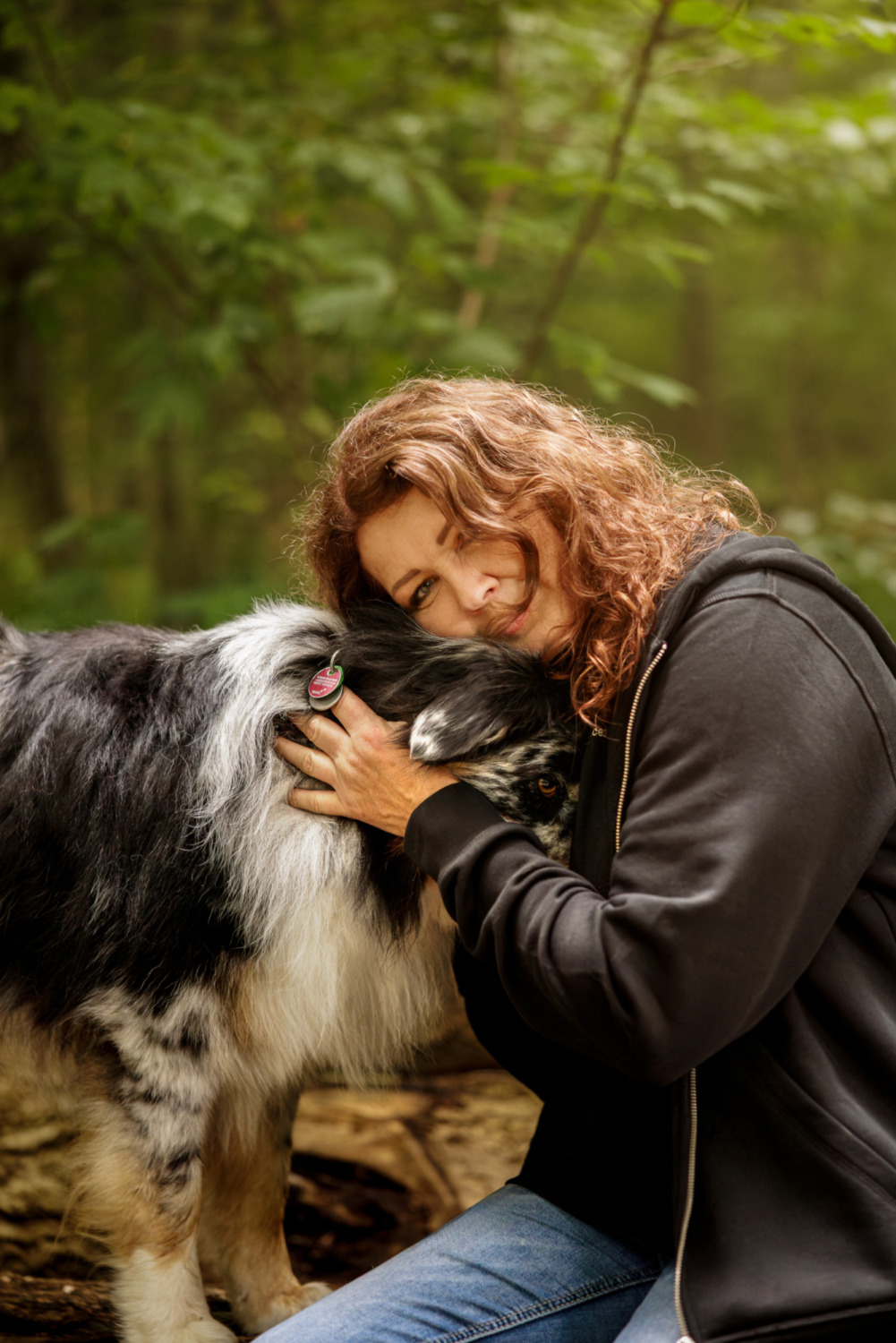 Frau mit langen rotbraunen Locken schmiegt ihr Gesicht an einen blue-merle Australian Shepherd