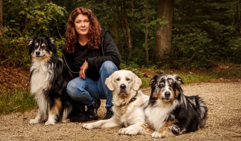 Frau kniet auf einem Waldweg, links ein Australian Shepherd, vor ihr liegen ein Golden Retriever und ein blue-merle Aussie