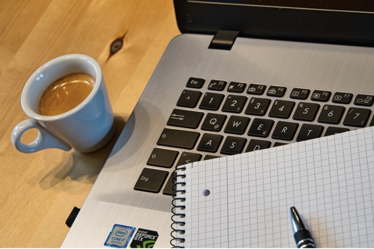 Laptop auf einem Holztisch, daneben eine hellblaue Espressotasse, ein kariertes Notizbuch und ein Kugelschreiber
